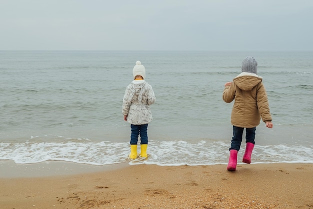 twee kleine meisjes in rubberen laarzen staan aan de kust in de herfst, lentezusjes spelen in de woestijn