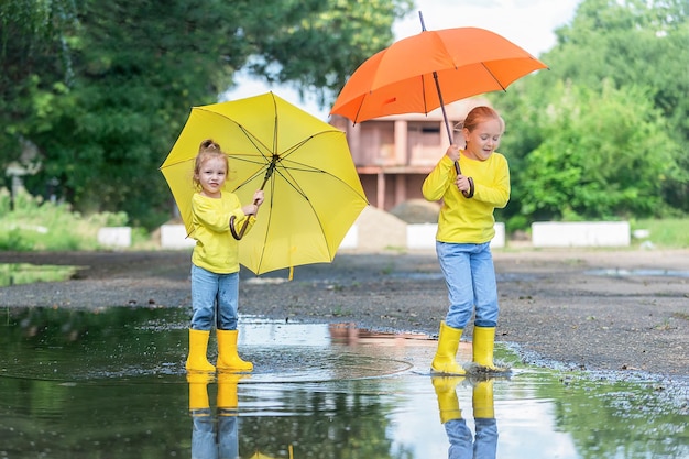 twee kleine meisjes lopen na de regen door de plassen in rubberen laarzen en onder gele paraplu's