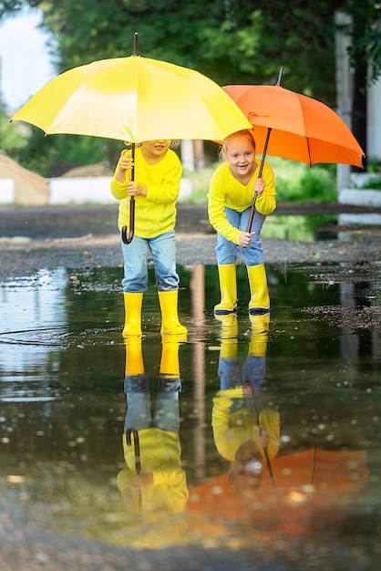 Twee kleine meisjes met felgele en oranje paraplu's rennen door de plassen in rubberen laarzen