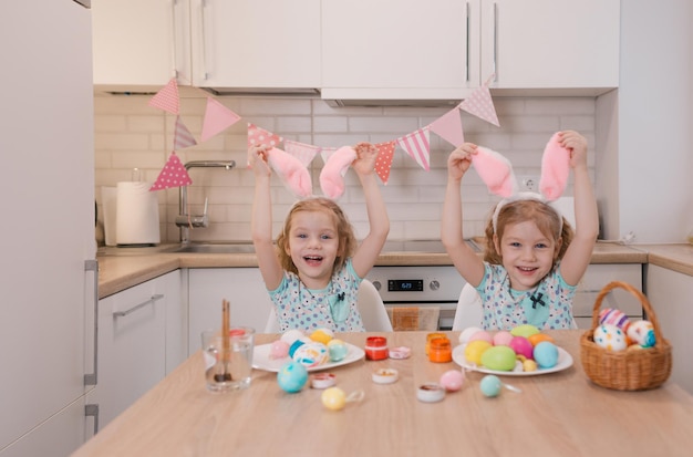 Twee kleine tweelingmeisjes raken met hun handen de konijnenoren op hun hoofd aan terwijl ze zich voorbereiden op de paasvakantie en lachen vrolijk