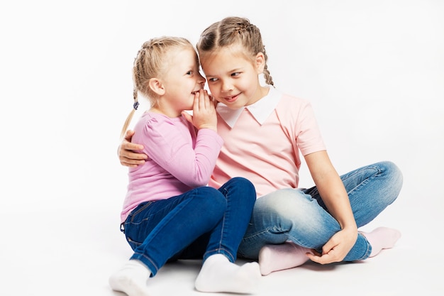 Twee kleine vriendinnen in jeans en roze truien roddelen. witte muur.