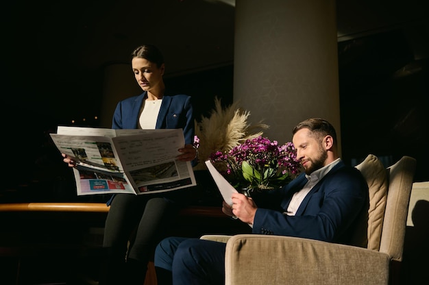 Twee knappe mensen, blanke man van middelbare leeftijd en mooie elegante vrouw in pak, collega's, zakenpartners die een krant lezen in de hotellobby