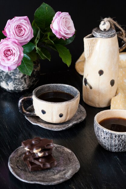 Twee kopjes zwarte koffie, zwarte chocolade, een gele theepot en een vaas met roze rozen.