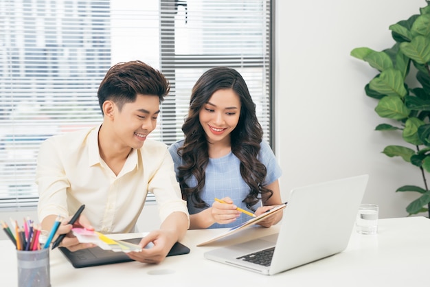 Twee lachende casual ontwerpers die met laptop werken terwijl ze aan een bureau op kantoor zitten