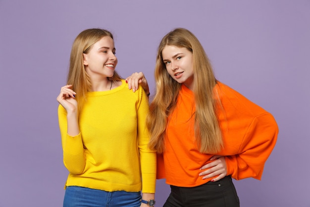 Twee lachende jonge blonde tweelingzusters meisjes in levendige kleurrijke kleding staan, opzij geïsoleerd op pastel violet blauwe muur. Mensen familie levensstijl concept.