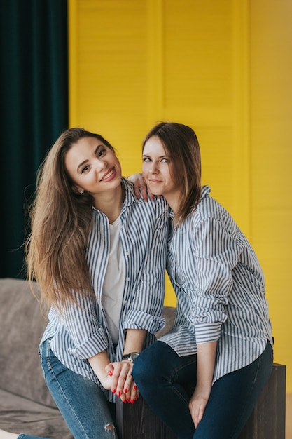 Foto twee lachende jonge meisjes in gestreepte shirts, jeans en sneakers die zich voordeed op de bank. studio-opnamen.