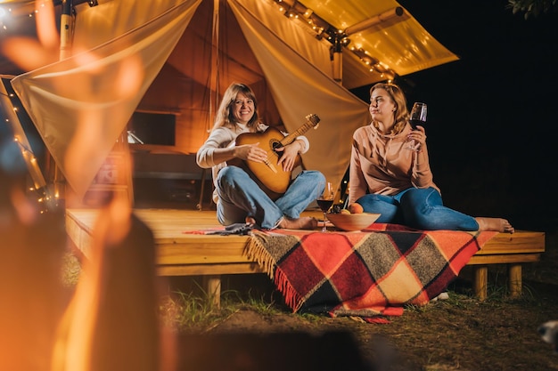 Twee lachende vriendinnen wijn drinken en gitaar spelen zitten in gezellige glamping tent in herfst avond vreugdevuur luxe camping tent voor buiten vakantie en vakantie Lifestyle concept