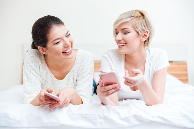Twee lachende vrouwen die thuis smartphones op bed gebruiken