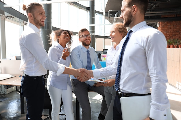 Twee lachende zakenlieden schudden handen terwijl ze samen met hun collega's op kantoor zijn.