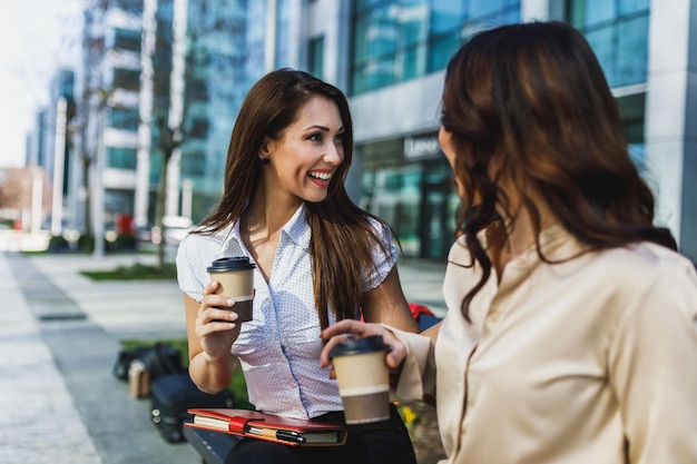 Twee lachende zakenvrouwen chatten tijdens de koffiepauze buiten een kantoor.