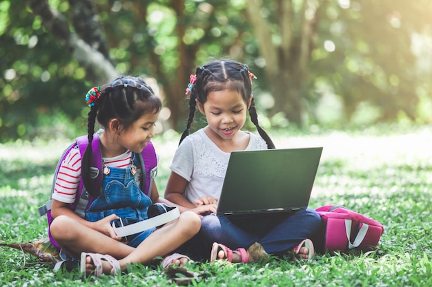 Twee leuke Aziatische kindmeisjes die laptop in het park samen met pret en geluk gebruiken