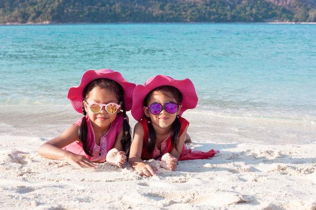 Twee leuke Aziatische kindmeisjes die roze hoed en zonnebril dragen die samen met zand op het strand spelen