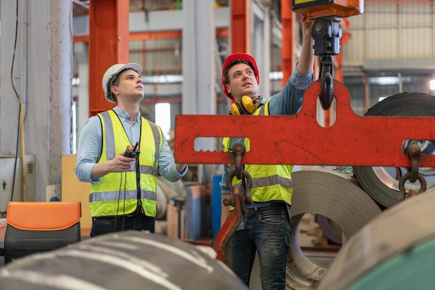 Twee mannelijke ingenieurs dragen een veiligheidsvest met helm die de bovenloopkraan van de fabriek controleert en bestuurt