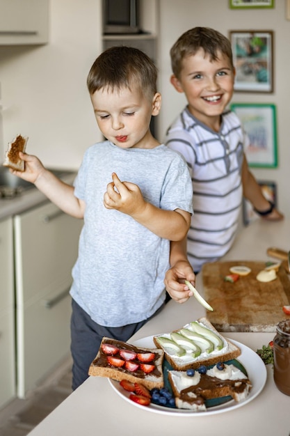 Twee mannelijke kleine vriendenbroer die de ochtendsandwich versieren met fruitbessen en chocoladepasta