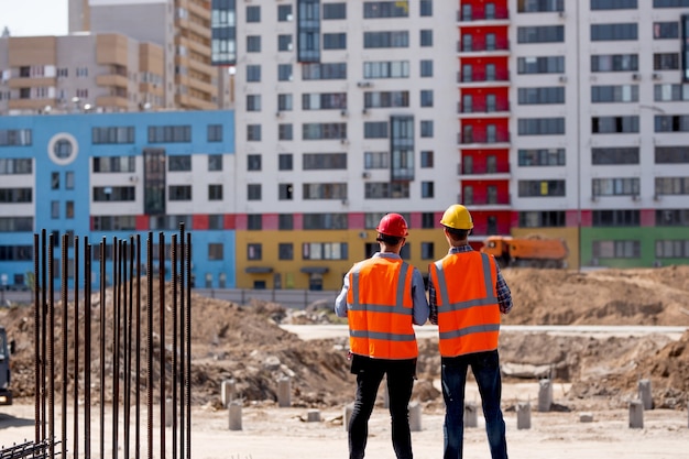 Twee mannen gekleed in oranje werkvesten en helmen vertellen over het bouwproces op de bouwplaats tegen de achtergrond van een gebouw met meerdere verdiepingen.
