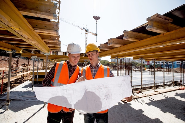 Twee mannen gekleed in overhemden, oranje werkvesten en helmen verkennen bouwdocumentatie op de bouwplaats bij de houten bouwconstructies.