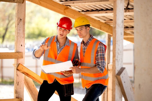 Twee mannen gekleed in overhemden, oranje werkvesten en helmen verkennen bouwdocumentatie op de bouwplaats bij de houten bouwconstructies.