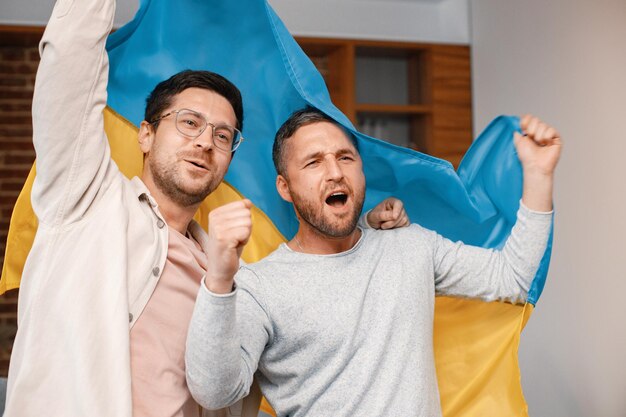 Twee mannen kijken naar een voetbalwedstrijd op tv en houden een grote vlag van Oekraïne vast