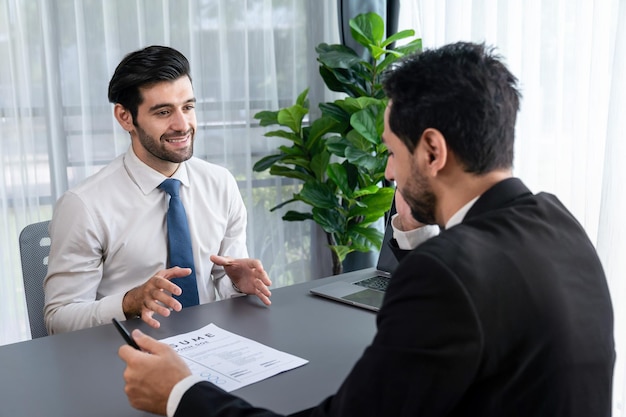 Twee mannen zitten aan een bureau, een van hen draagt een blauwe stropdas en de ander is in gesprek met een andere man.