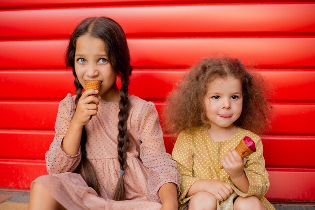 Twee meisjes eten ijs bij het rode hek. Een donkerbruin meisje met twee staartjes, de tweede licht krullende krul