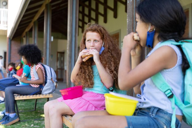 Twee meisjes lunchen uit een tiffin-doos terwijl ze op een bankje in het park op school zitten