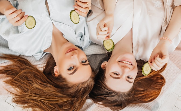 Twee meisjes maken zelfgemaakte gezichts- en haarschoonheidsmaskers. Komkommers voor de frisheid van de huid rond de ogen. Vrouwen zorgen voor een jeugdige huid. Vriendinnen lachen thuis liggend op de vloer op kussens.