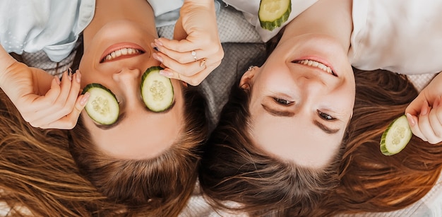 Twee meisjes maken zelfgemaakte gezichts- en haarschoonheidsmaskers. Komkommers voor de frisheid van de huid rond de ogen. Vrouwen zorgen voor een jeugdige huid. Vriendinnen lachen thuis liggend op de vloer op kussens.