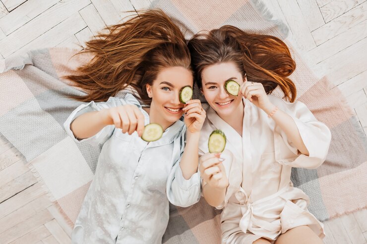 Twee meisjes maken zelfgemaakte gezichts- en haarschoonheidsmaskers. Komkommers voor de frisheid van de huid rond de ogen. Vrouwen zorgen voor een jeugdige huid. Vriendinnen lachen thuis liggend op de vloer op kussens.