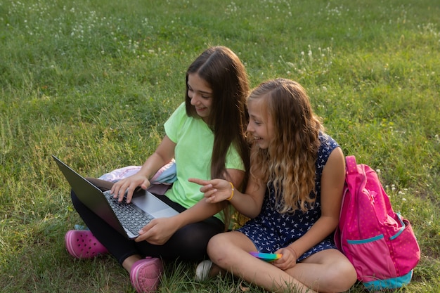 Twee meisjes met laptop en aktetassen die huiswerk maken of plezier hebben terwijl ze buiten op het gras zitten