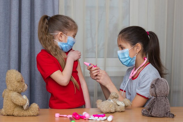 Twee meisjes met medische maskers spelen met speelgoed in het ziekenhuis, het jongste meisje dat wordt gevaccineerd