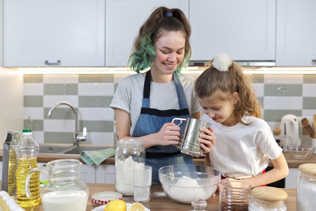 Twee meisjes, tiener en jongere zus, bereiden samen koekjes in de keuken. Kinderen roeren meel, de oudste laat de jongste zien. Familie, vriendschap, plezier, gezond zelfgemaakt eten