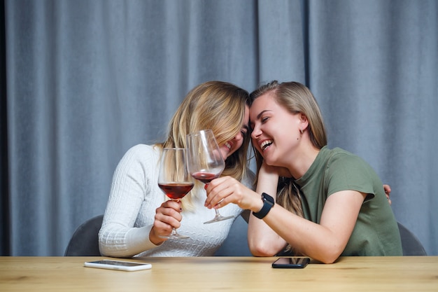 Twee meisjes van Europees uiterlijk met blond haar zitten aan tafel, drinken wijn en lachen, ontspannen thuis, alcohol