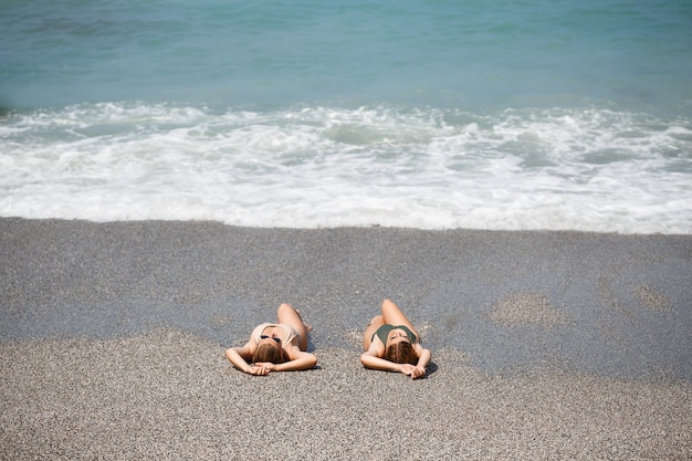 Twee meisjes vriendin zitten op de zanderige kust van de zee en de golven doordrenkt hen in badpakken op een zonnige warme dag