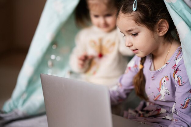 Twee meisjeszusters die op laptop bij wigwamtent letten Technologie en huisconcept