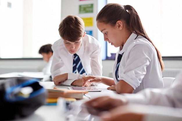 Twee middelbare scholieren in uniform werken samen aan bureau met behulp van digitale tablet
