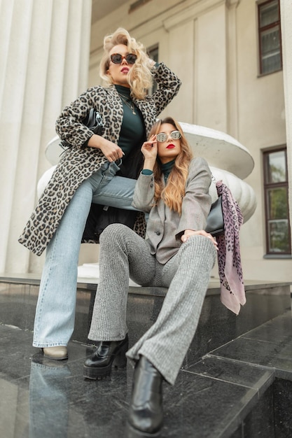 Twee mode jonge mooie vrouwen met trendy zonnebril in modieuze kleding met elegante pak luipaard jas en schoenen zit en poseert in de buurt van een vintage gebouw
