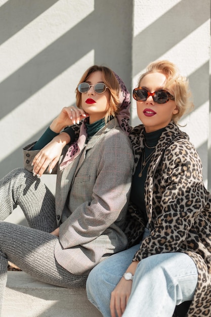 Twee mode mooie jonge meisjes met vintage zonnebril in modieuze outfit met luipaard jas spijkerbroek en grijs pak zit en poseert op straat in de buurt van muur in zonlicht vrouwelijke stedelijke stijl kleding