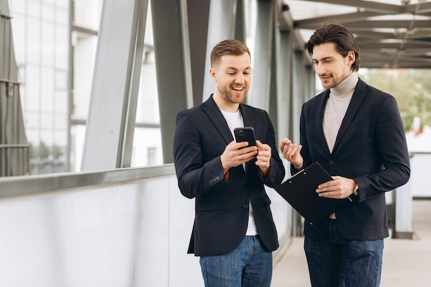 Twee moderne zakenlieden bespreken iets en tonen elkaar op een smartphone tegen de achtergrond van stedelijke kantoren en gebouwen