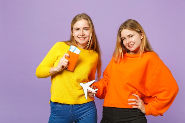 Twee mooie blonde tweelingzusjes meisjes in kleurrijke kleding houden paspoort instapkaart ticket, papieren vliegtuigje geïsoleerd op violet blauwe muur. Mensen familie levensstijl concept.