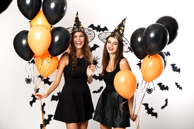 Twee mooie brunette meisjes gekleed in zwarte jurken en hoeden houden zwarte en oranje ballonnen vast. halloween.