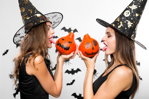 Twee mooie brunette meisjes in zwarte heksenhoeden houden halloween-pompoenen in hun handen en tonen hun tong.