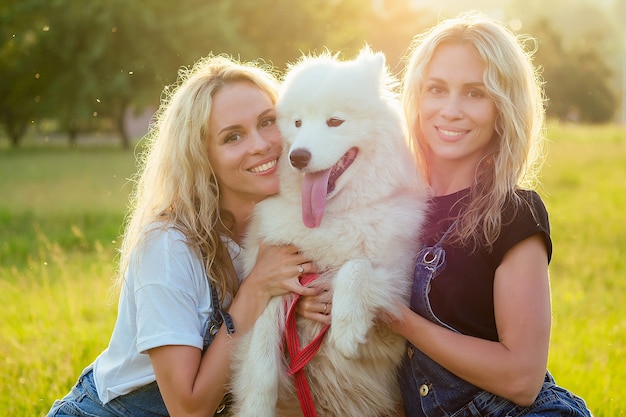 Twee mooie en charmante krullende blonde tweelingen glimlachende toothy vrouw in denim overalls zitten met een witte pluizige Samojeed hond, in het zomerpark bij glazen zonsondergang stralen achtergrond.