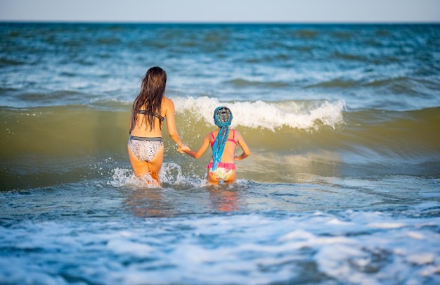 Twee mooie gebruinde langharige vrolijke zussen in heldere zomerbadpakken krimpen ineen en springen in blauwe schuimige zeegolven op een zandige ondiepte met kleine schelpen, op een warme zonnige avond van vakantie