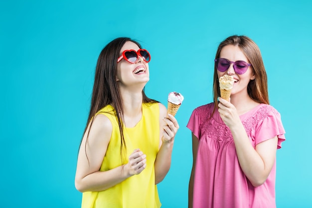 Twee mooie gelukkige lachende meisjes gekleed in kleurrijke t-shirts en zonnebrillen hebben plezier met ijs op blauwe achtergrond zorgeloos jeugdconcept