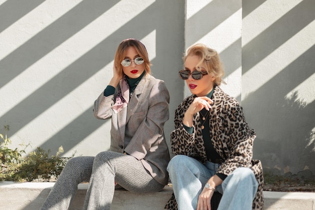 Twee mooie glamoureuze jonge vriendinnen vrouwen met retro zonnebril in stijlvolle outfit met luipaard jas pak spijkerbroek zit op straat in zonlicht lijn Cool vintage mooie meisjes buitenshuis