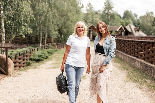 Twee mooie glimlachende vrouwen, moeder en gelukkige dochter in een wit poloshirt met blauwe spijkerbroek en een jas lopen buiten