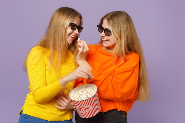 Twee mooie jonge blonde tweelingzusters meisjes in 3d imax-bril kijken naar filmfilm, met popcorn geïsoleerd op pastel violet blauwe muur. Mensen familie levensstijl concept.