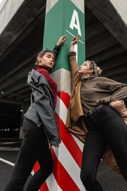 Twee mooie jonge modelmeisjes in modieuze kleding met zwarte jeans en een stijlvol jasje poseren op een parkeerplaats in de stad