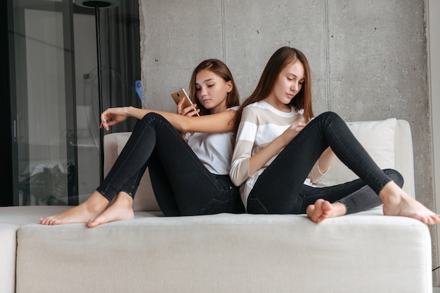 Twee mooie jonge tienermeisjes met behulp van mobiele telefoons zittend op een bank thuis