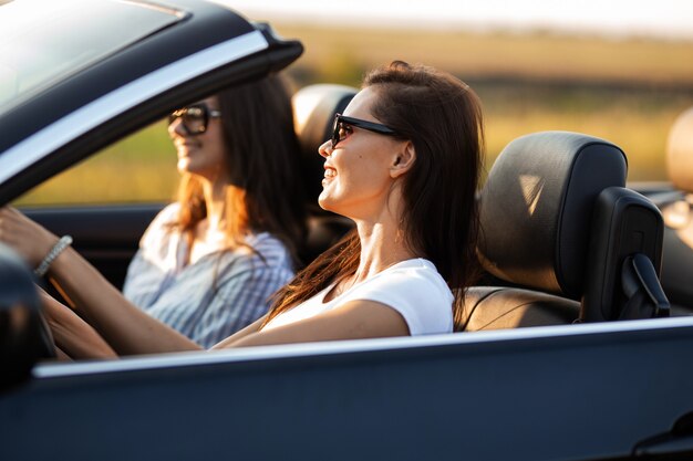 Twee mooie jonge vrouwen in zonnebril zitten in een zwarte cabriolet en glimlachen op een zonnige dag. .